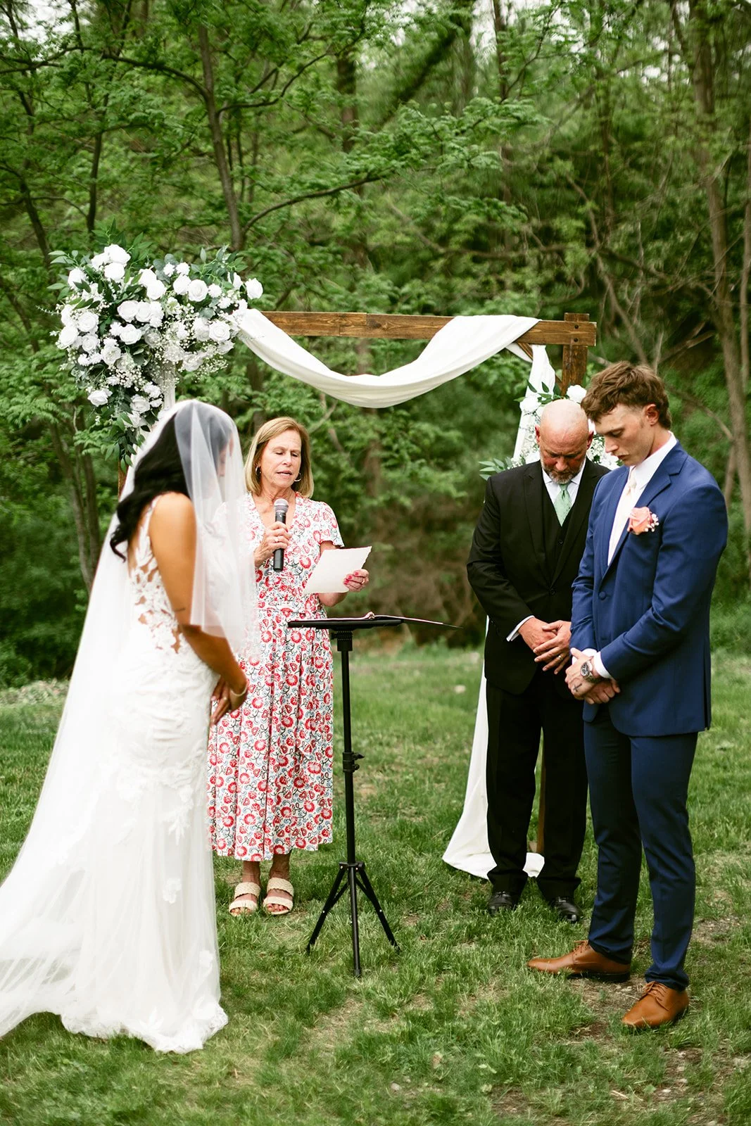 A bride and groom stand with heads bowed beside an officiant and another man under a wooden wedding arch with white flowers and drapery, outdoors.