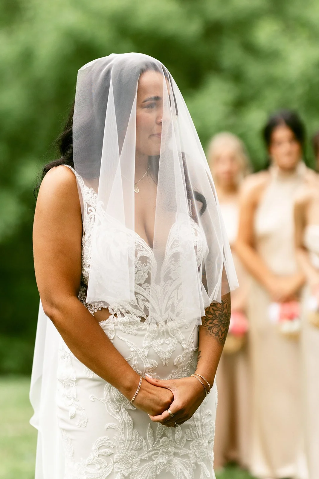 A bride and groom stand with heads bowed beside an officiant and another man under a wooden wedding arch with white flowers and drapery, outdoors.