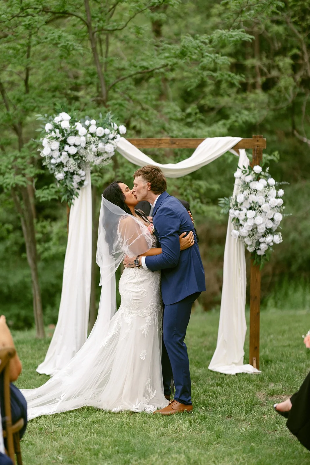 A bride and groom stand with heads bowed beside an officiant and another man under a wooden wedding arch with white flowers and drapery, outdoors.