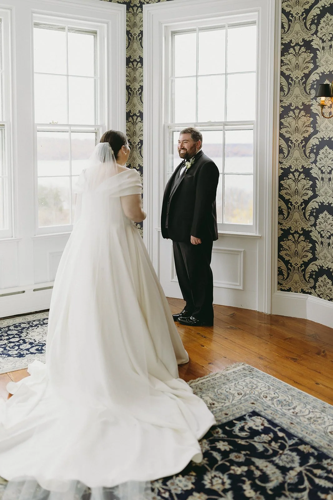 A bride in a white gown and a groom in a black suit stand facing each other in a room with large windows and patterned wallpaper.