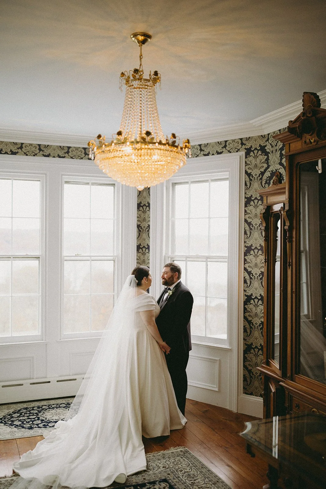 A bride in a white gown and a groom in a black suit stand facing each other in a room with large windows and patterned wallpaper.