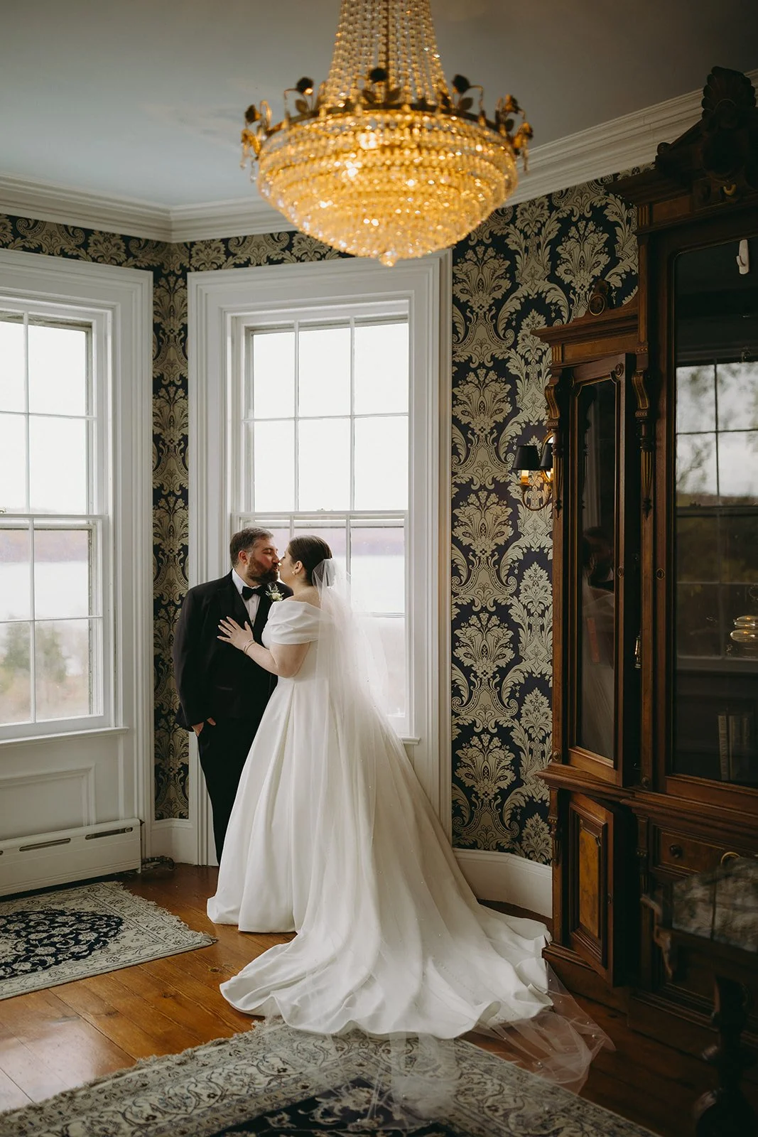 A bride in a white gown and a groom in a black suit stand facing each other in a room with large windows and patterned wallpaper.