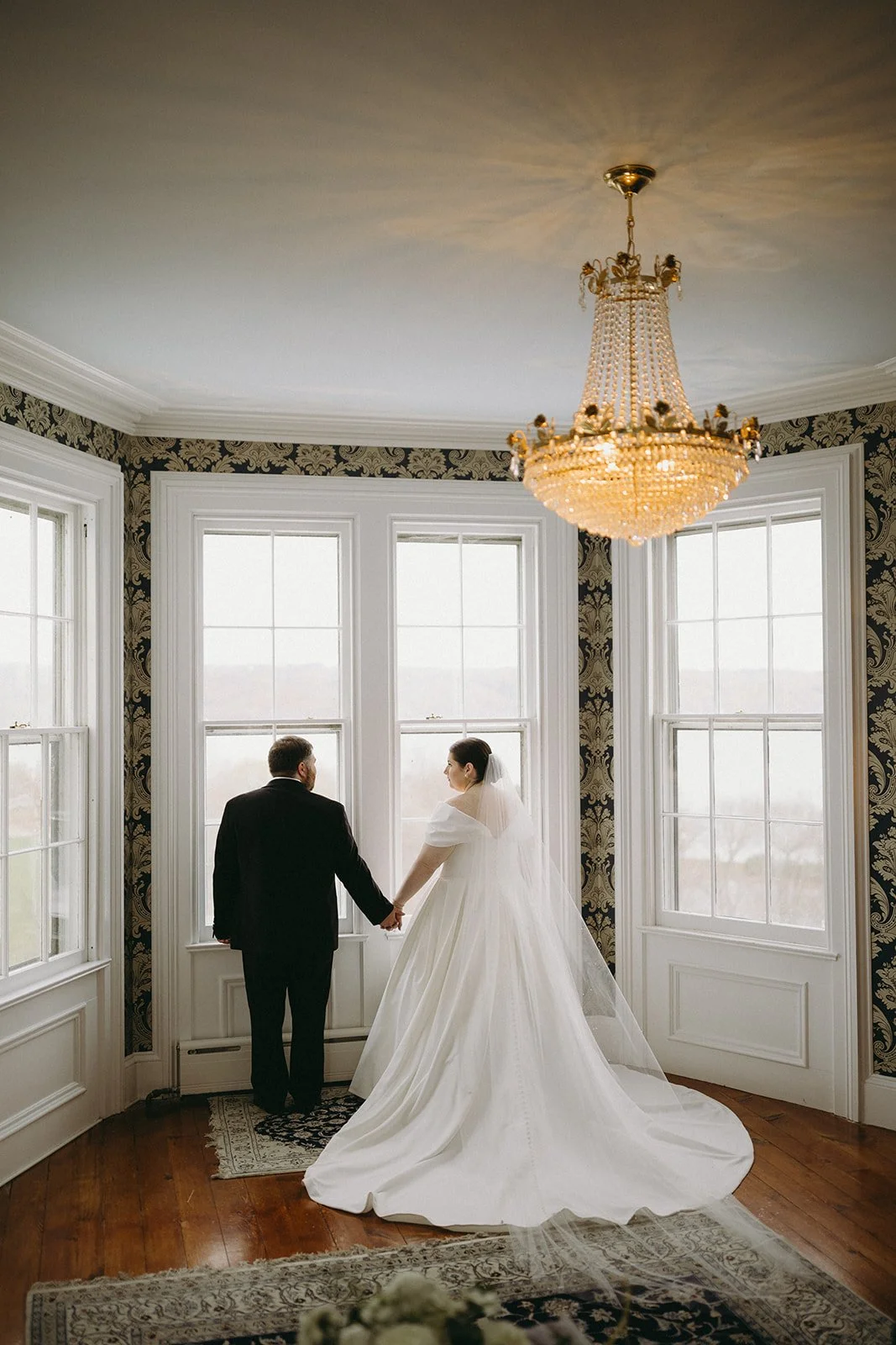 A bride in a white gown and a groom in a black suit stand facing each other in a room with large windows and patterned wallpaper.