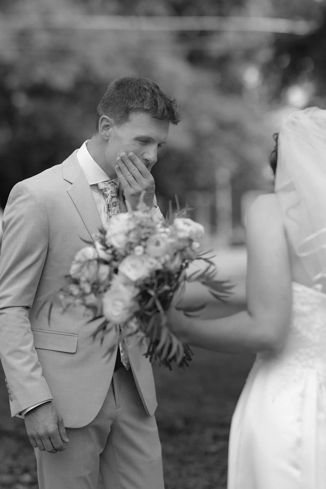 A groom in a light suit covers his mouth in surprise as he sees a bride in a wedding dress holding a bouquet outdoors.