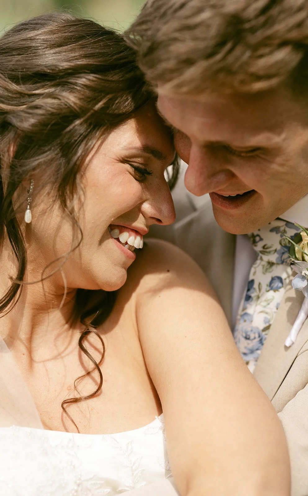 Bride and groom smiling closely at each other, both wearing formal attire, with the bride in a white dress and the groom in a light suit and floral tie.