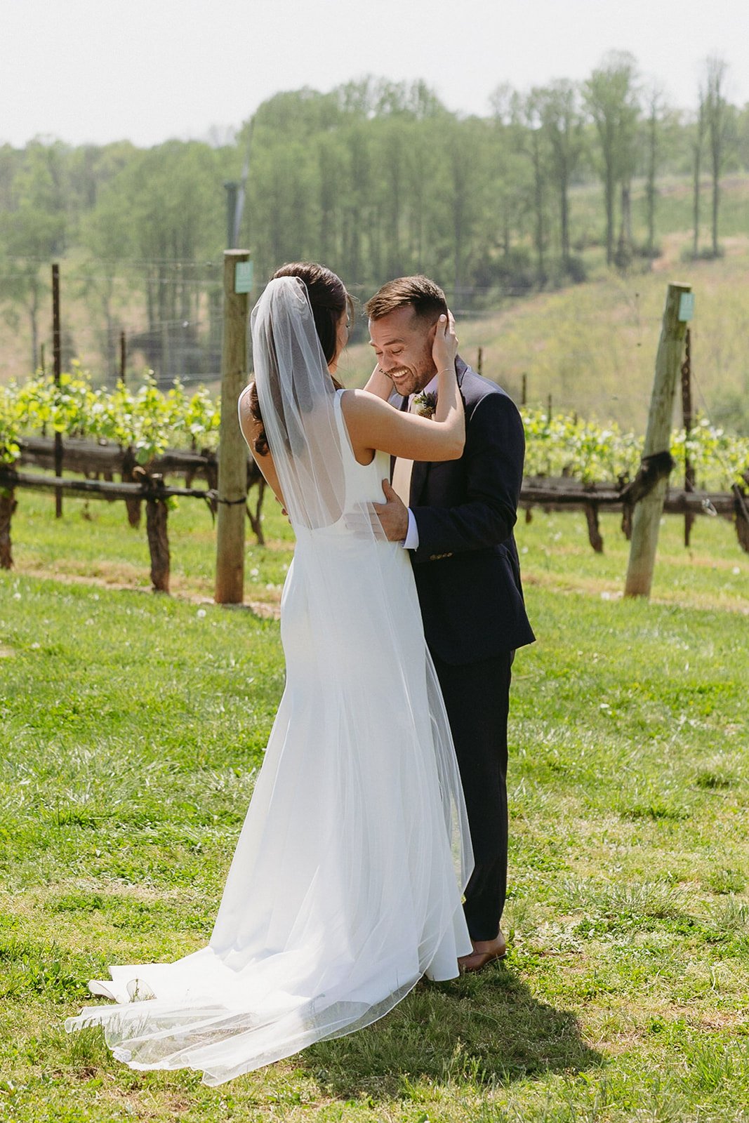 A bride in a white dress and veil approaches a groom in a dark suit from behind in an outdoor vineyard setting.
