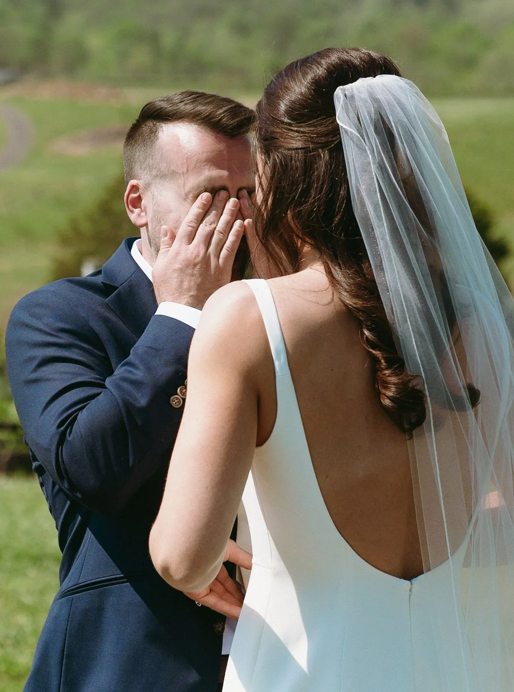 A bride and groom hold hands, walking down a grassy path between vineyard rows on a sunny day.