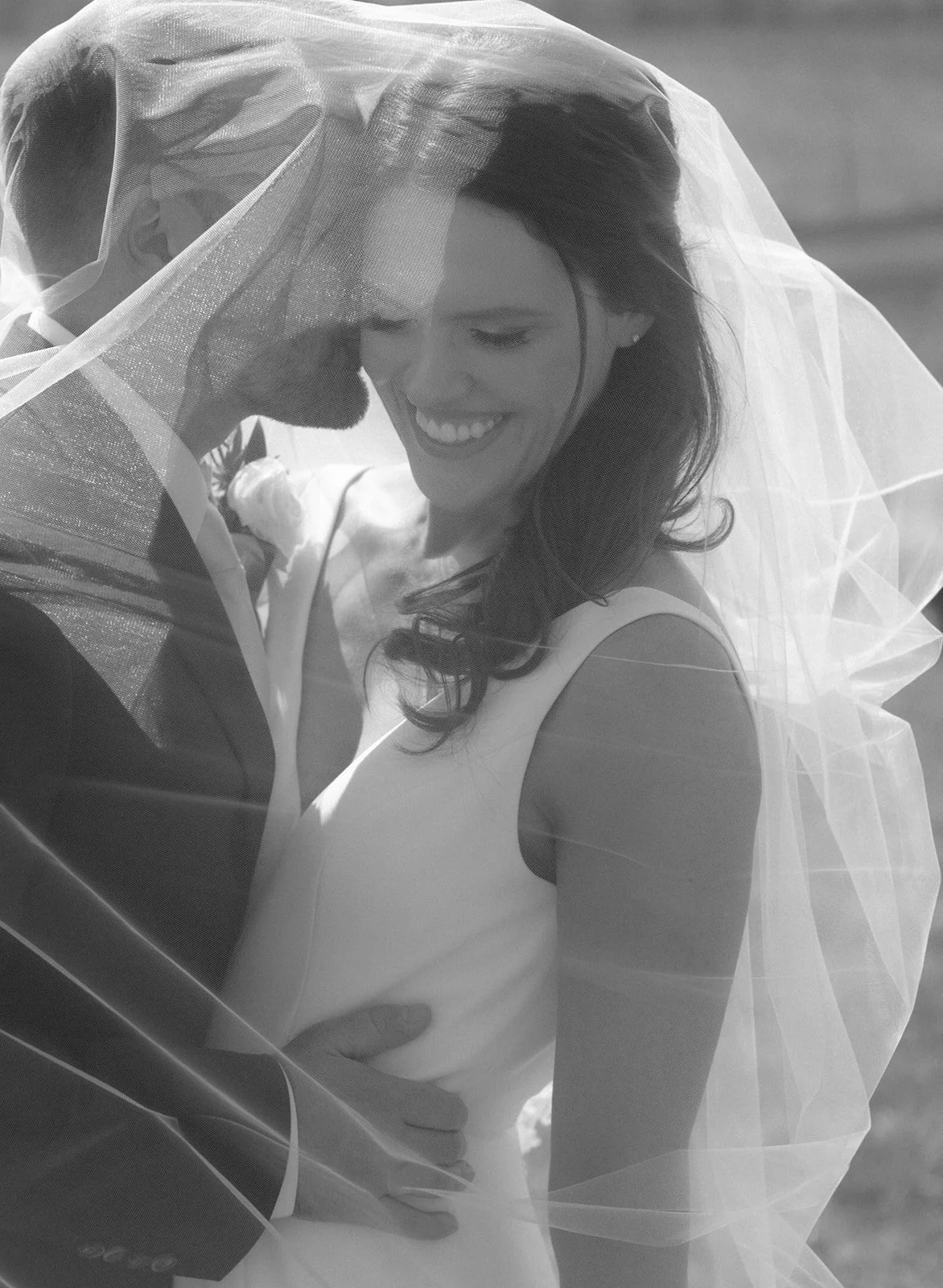 A bride and groom stand close together, smiling under a sheer wedding veil on their wedding day.