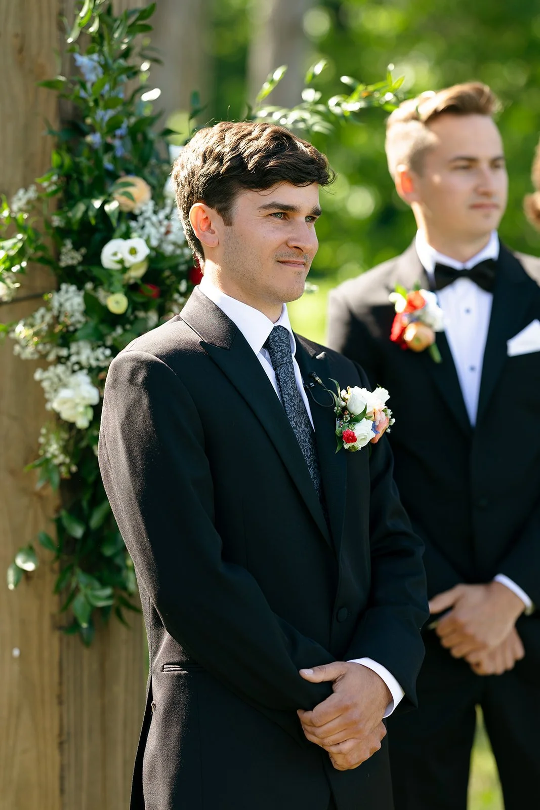 Two men in suits stand outdoors beside a wooden post decorated with flowers. The man in front wears a black suit and tie, and the man behind him wears a tuxedo.