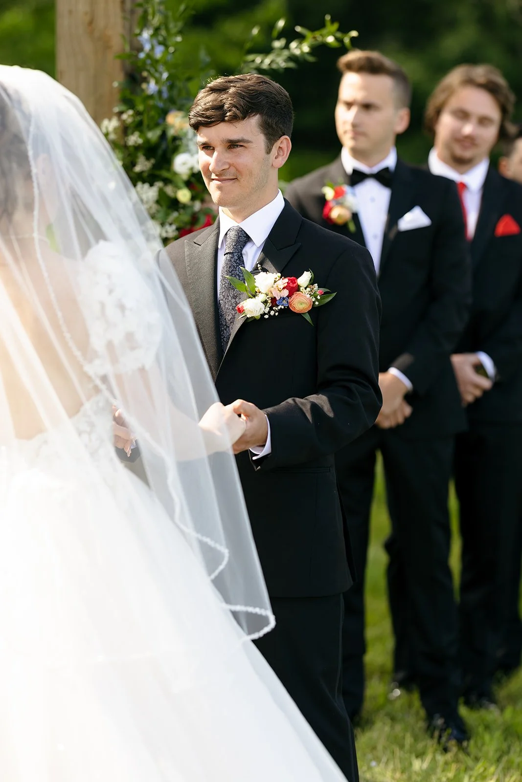A groom holds hands with a bride during an outdoor wedding ceremony, with groomsmen in black suits standing in the background.