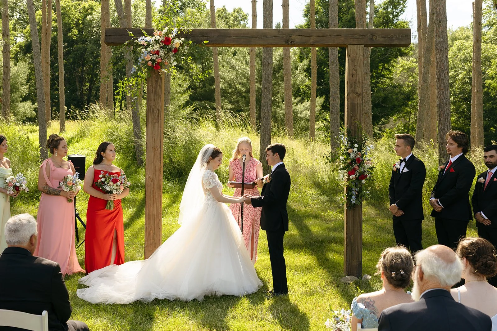 A bride and groom stand under a wooden arch exchanging vows outdoors, with six attendants beside them and guests seated in the foreground.