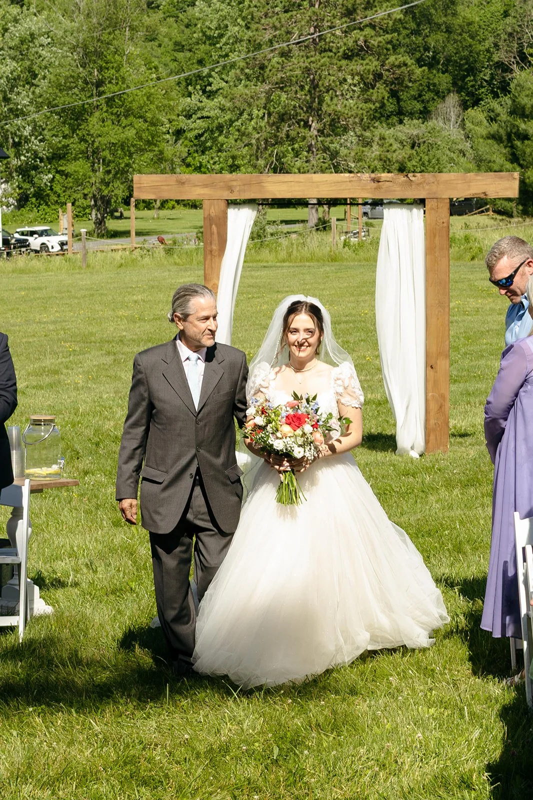 A bride in a white dress holding a bouquet walks down an outdoor aisle with an older man, as guests watch. A wooden arch with white fabric is in the background.