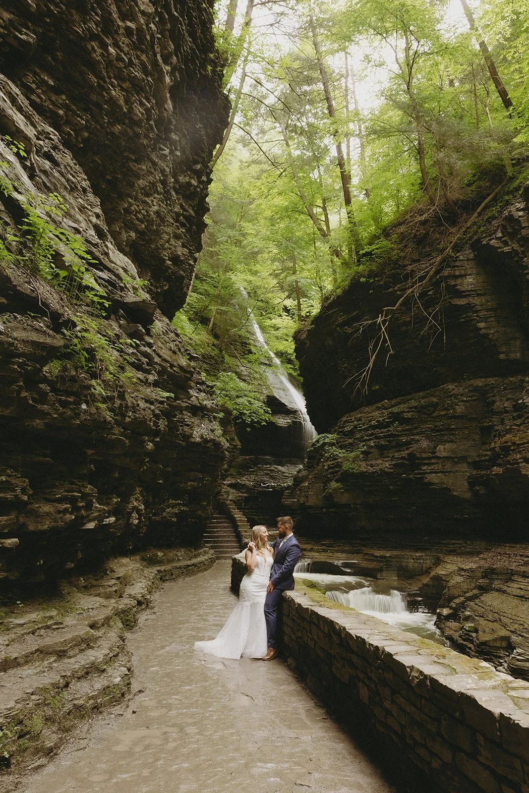 A couple in wedding attire stands by a stone wall in a rocky canyon with a waterfall and lush greenery in the background for Watkins Glen State Park Wedding Photos