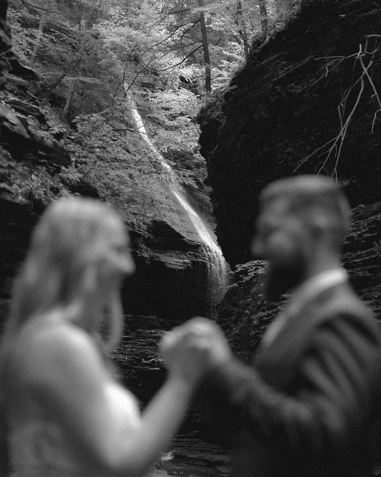 A couple in wedding attire stands by a stone wall in a rocky canyon with a waterfall and lush greenery in the background for Watkins Glen State Park Wedding Photos