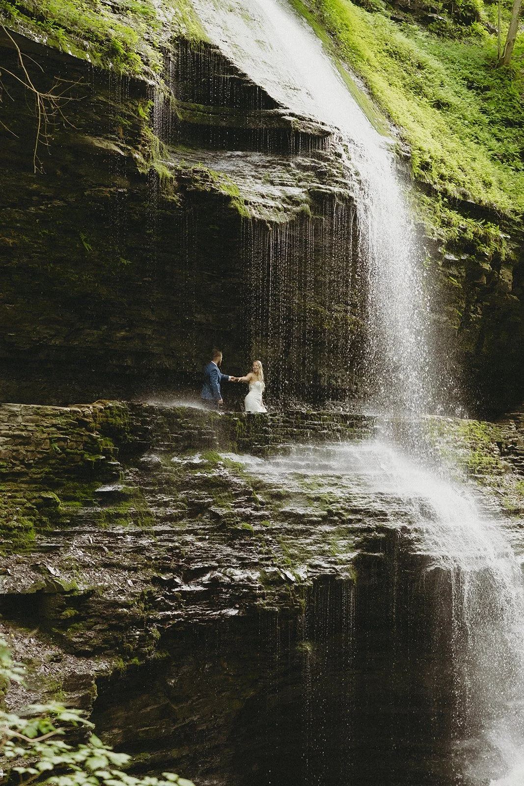 A couple in wedding attire stands by a stone wall in a rocky canyon with a waterfall and lush greenery in the background for Watkins Glen State Park Wedding Photos