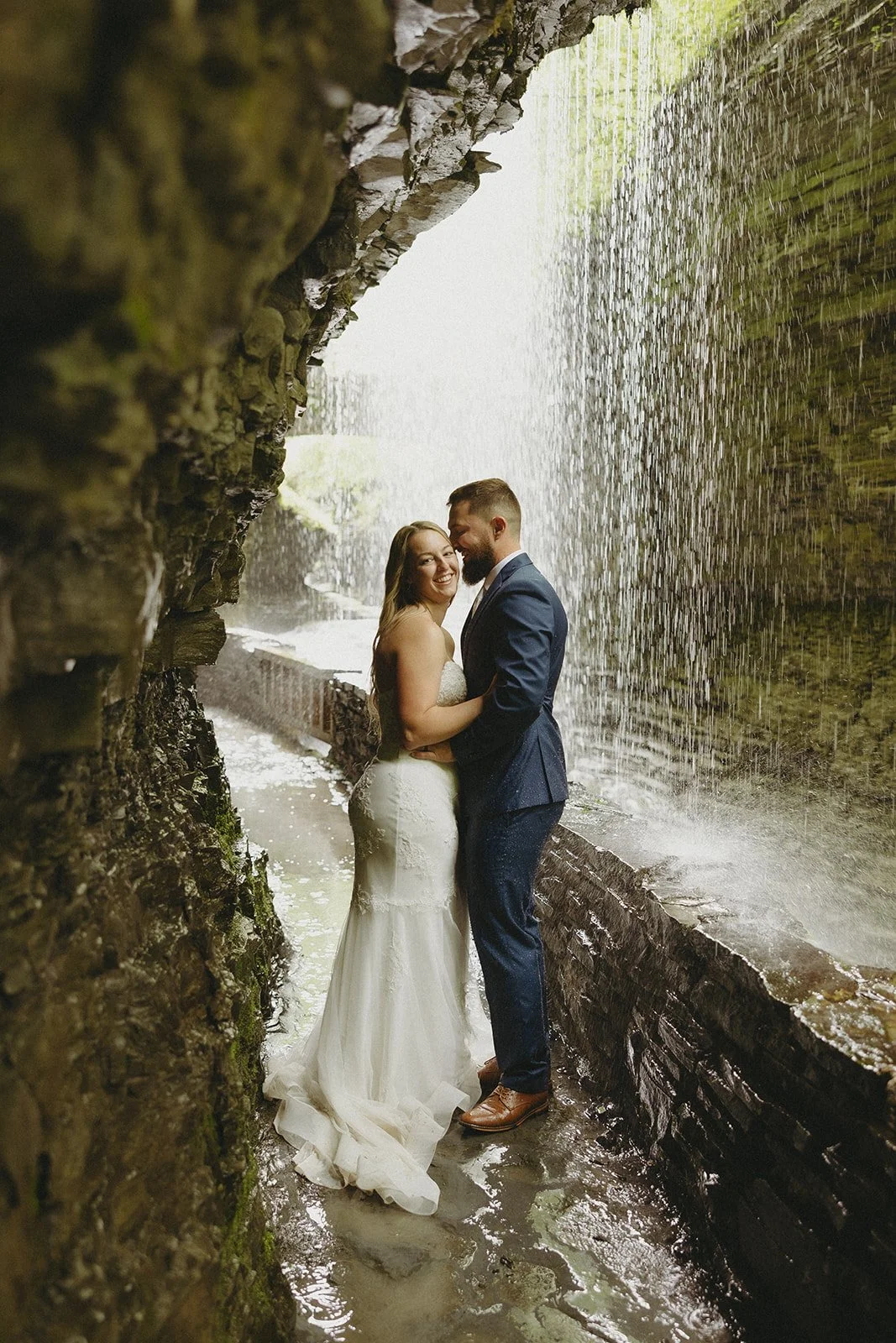A couple in wedding attire stands by a stone wall in a rocky canyon with a waterfall and lush greenery in the background for Watkins Glen State Park Wedding Photos