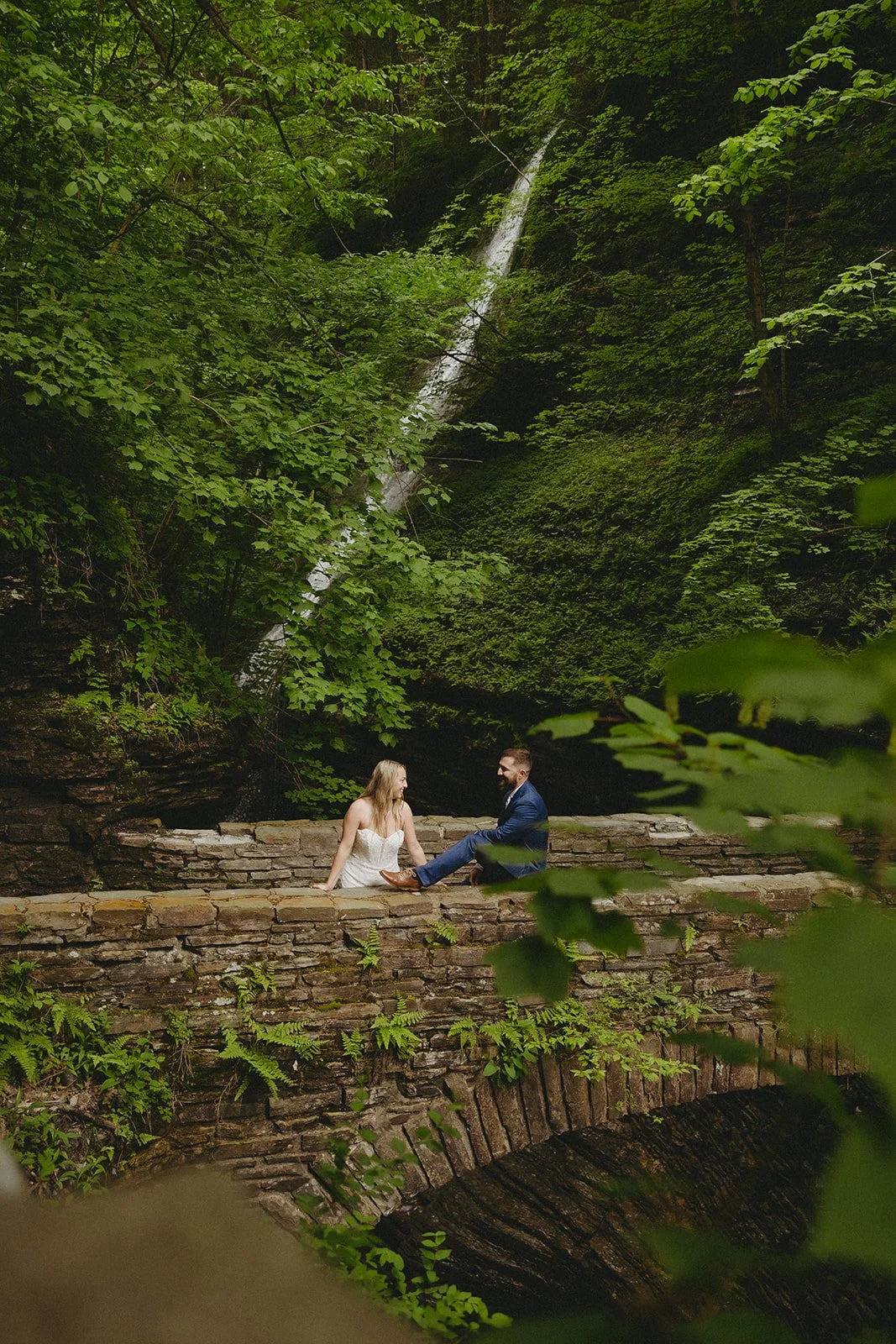 A couple in wedding attire stands by a stone wall in a rocky canyon with a waterfall and lush greenery in the background for Watkins Glen State Park Wedding Photos