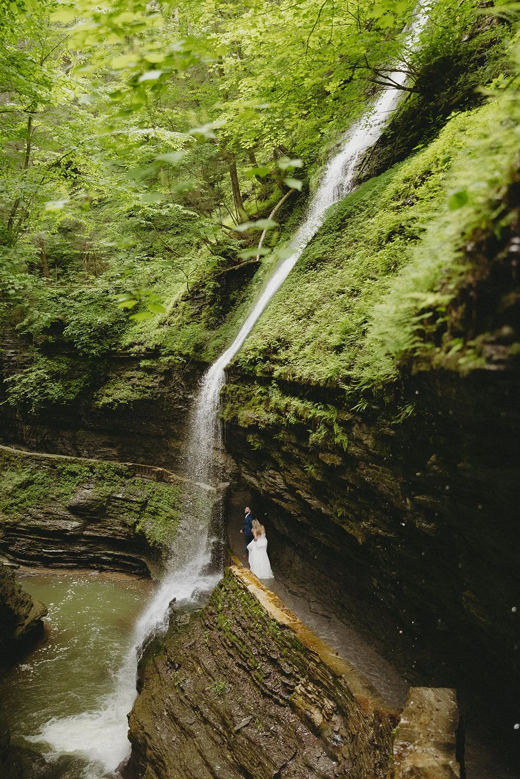 A couple in wedding attire stands by a stone wall in a rocky canyon with a waterfall and lush greenery in the background for Watkins Glen State Park Wedding Photos