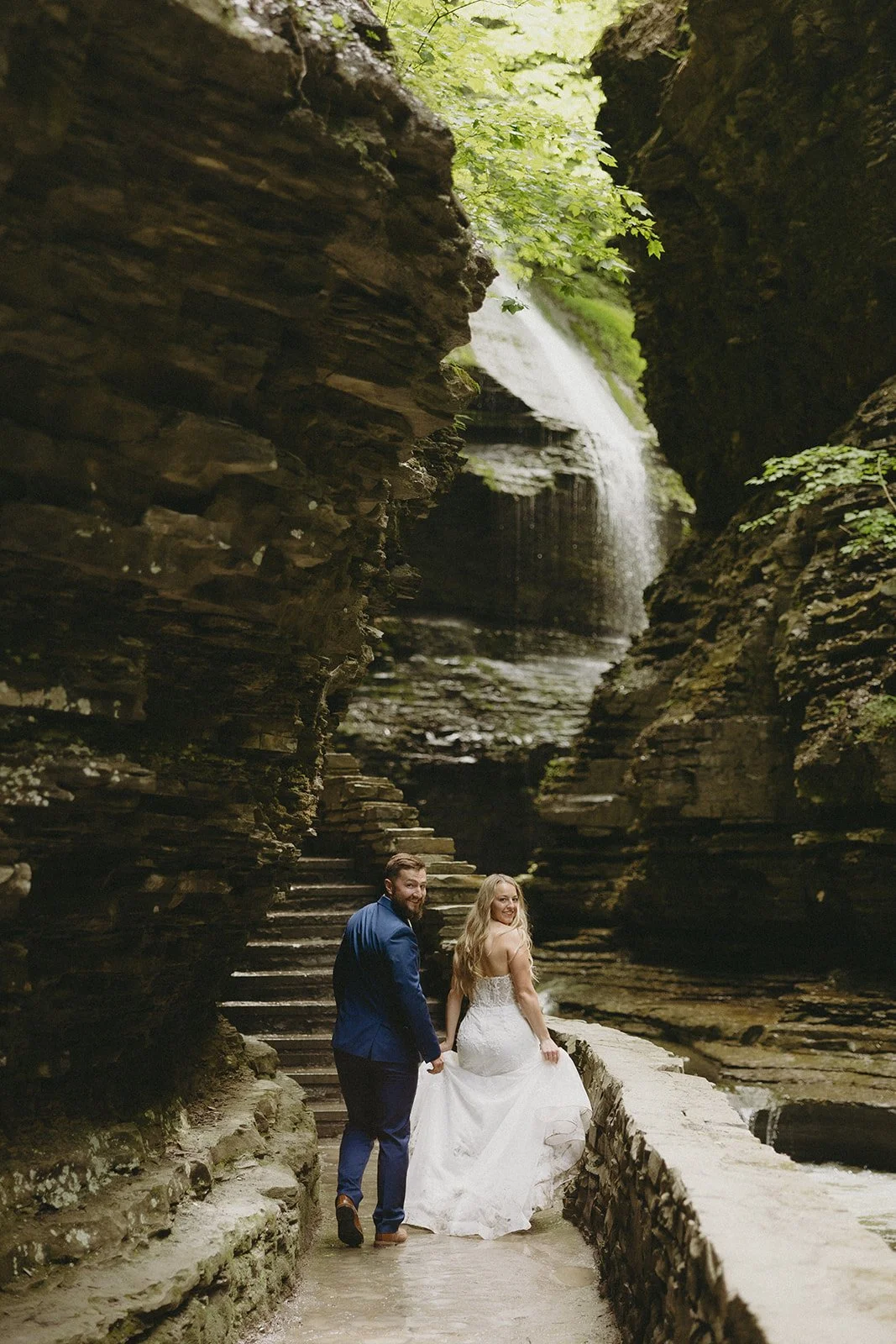 A couple in wedding attire stands by a stone wall in a rocky canyon with a waterfall and lush greenery in the background for Watkins Glen State Park Wedding Photos
