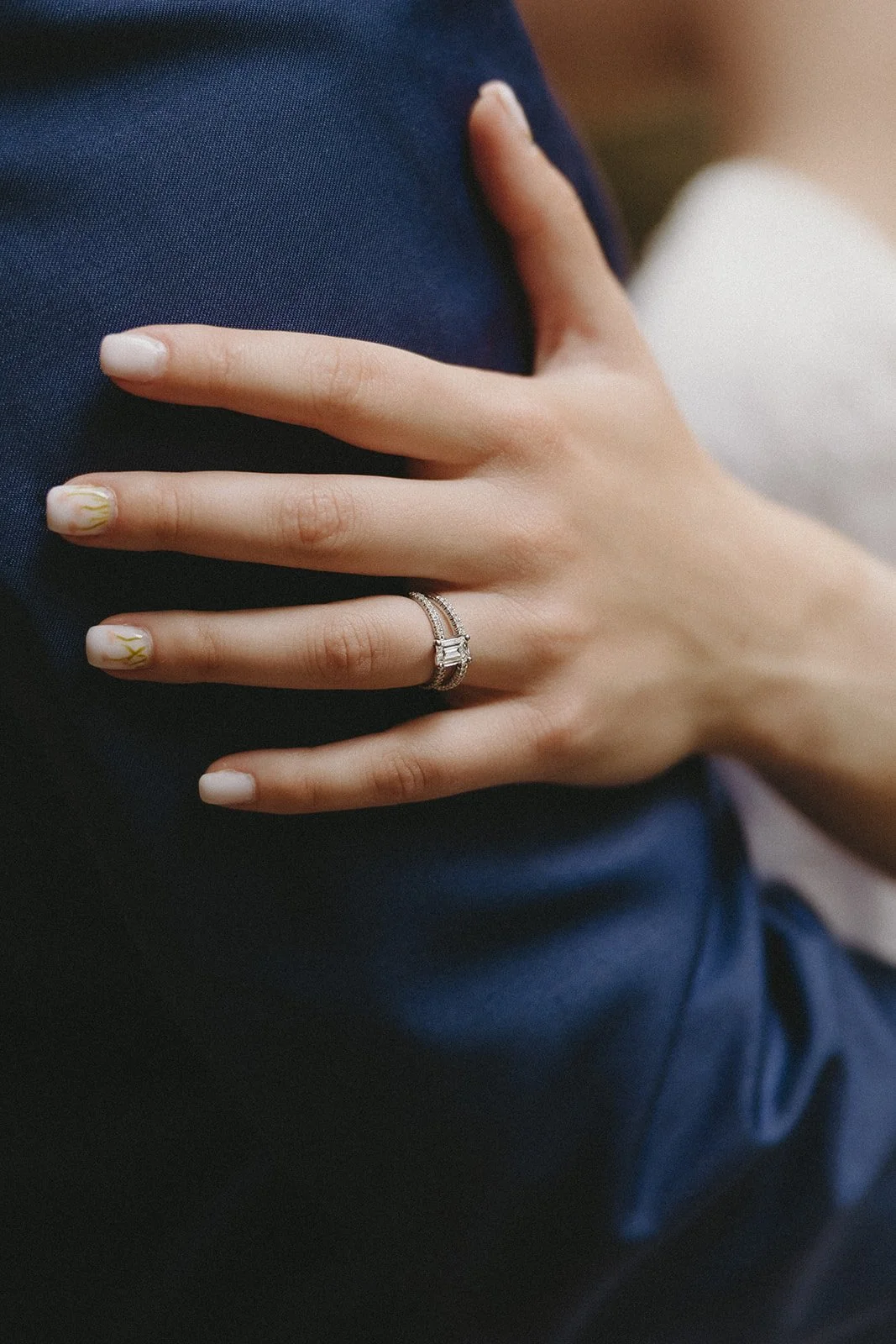A hand with a diamond engagement ring rests on someone wearing a dark blue suit.