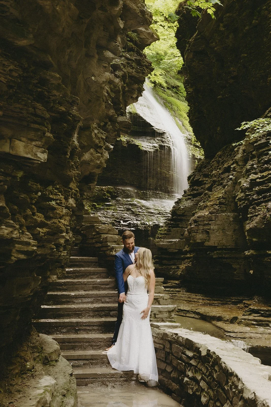 A couple in wedding attire stands by a stone wall in a rocky canyon with a waterfall and lush greenery in the background for Watkins Glen State Park Wedding Photos