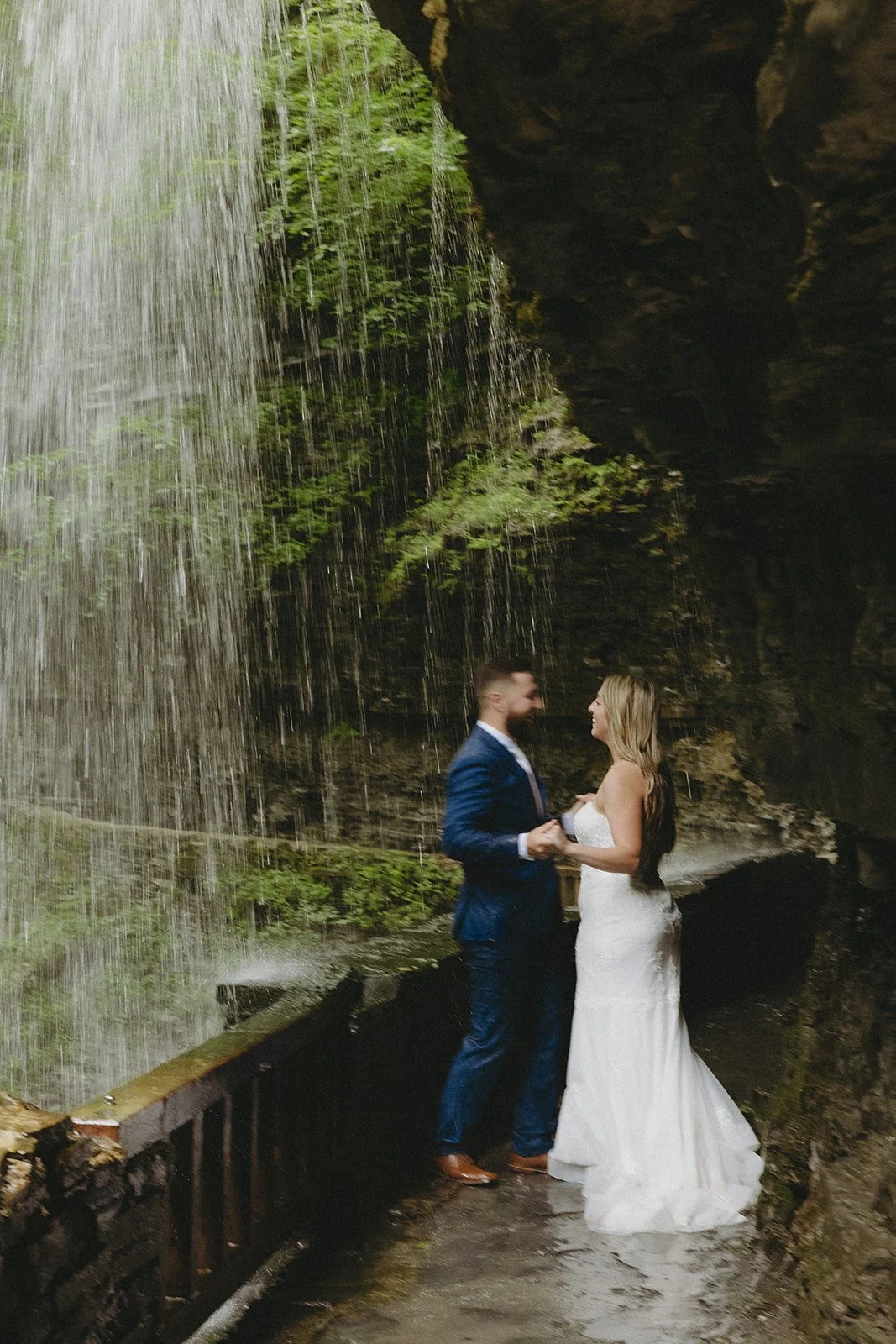 A couple in wedding attire stands by a stone wall in a rocky canyon with a waterfall and lush greenery in the background for Watkins Glen State Park Wedding Photos