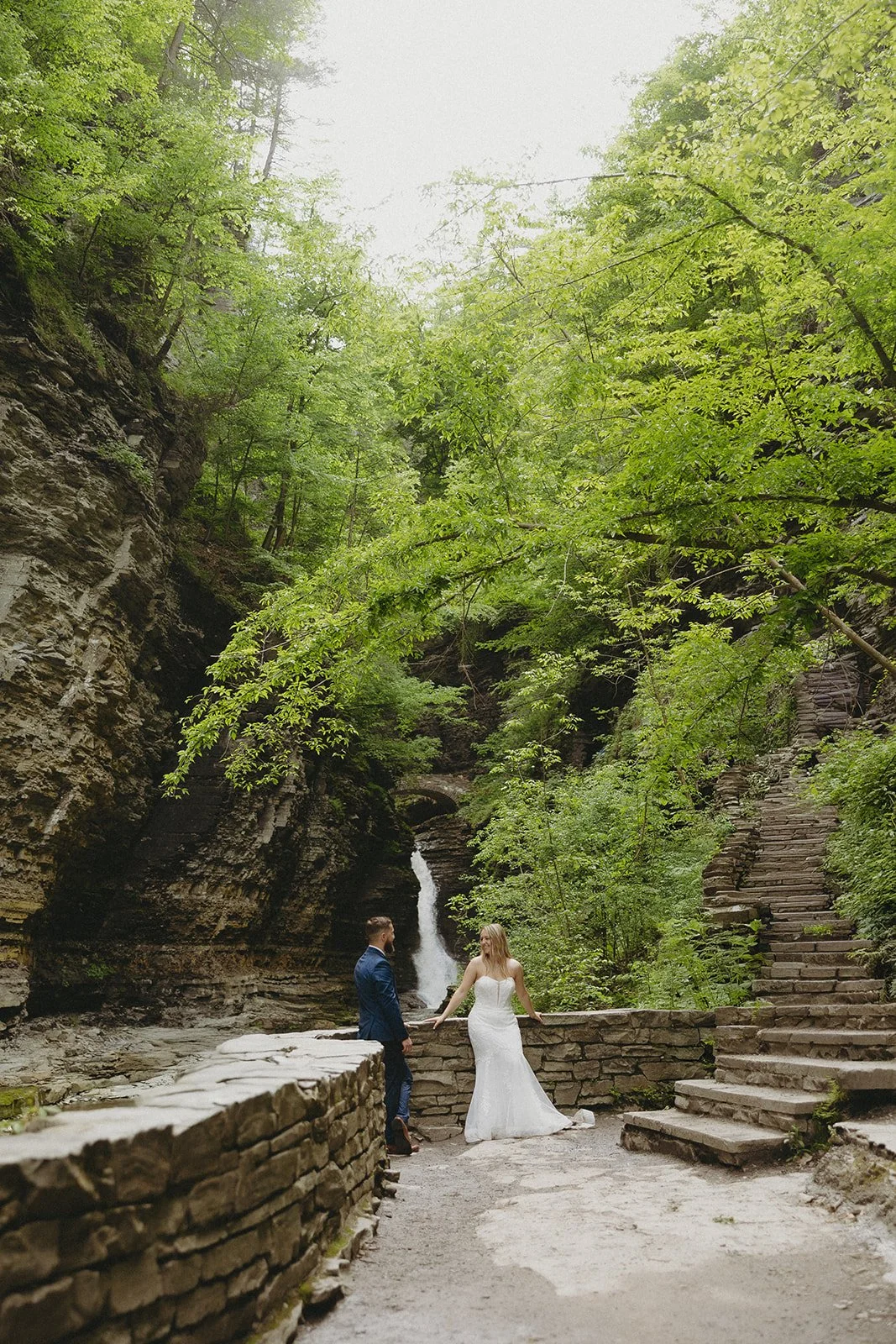 A couple in wedding attire stands by a stone wall in a rocky canyon with a waterfall and lush greenery in the background for Watkins Glen State Park Wedding Photos