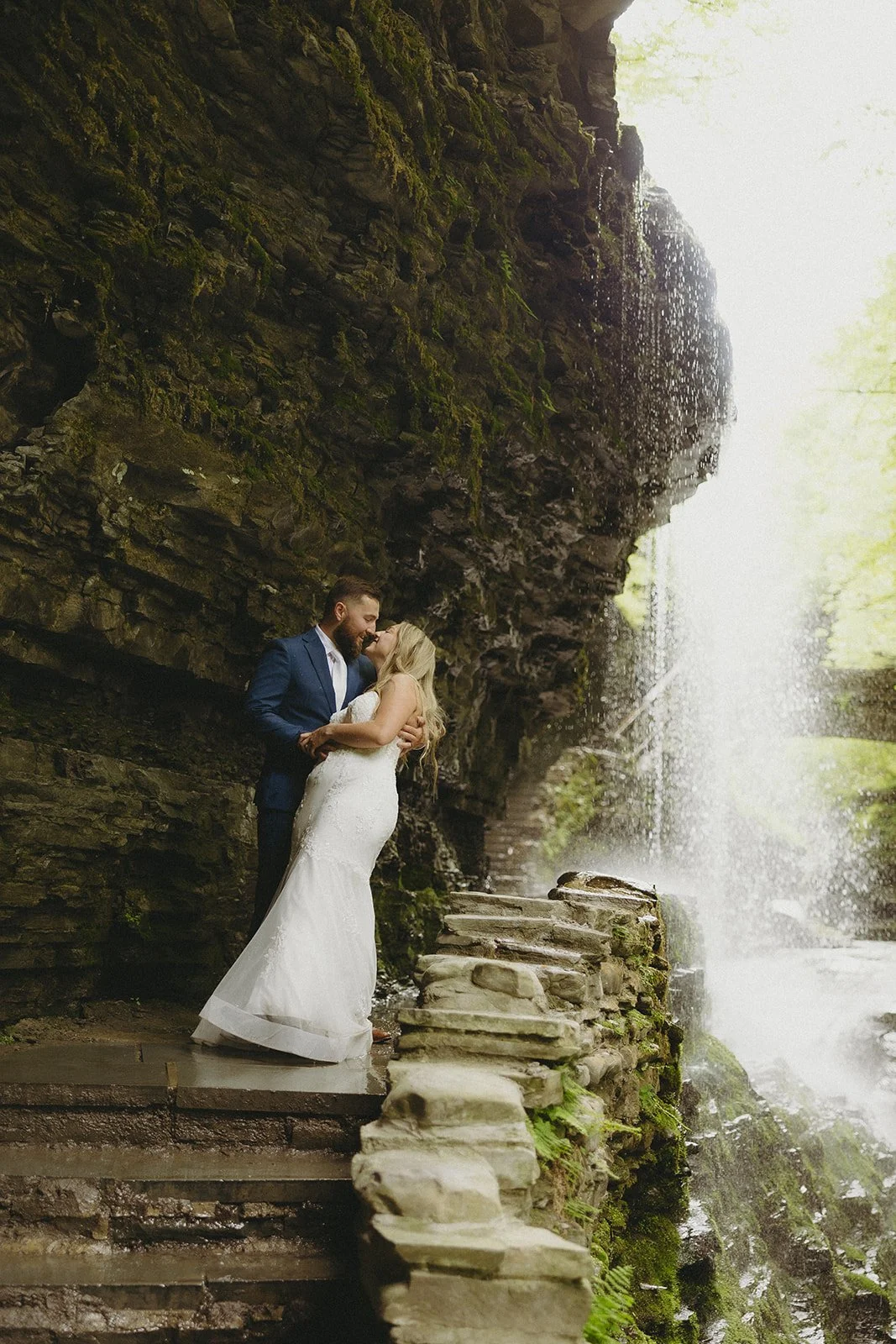 A couple in wedding attire stands by a stone wall in a rocky canyon with a waterfall and lush greenery in the background for Watkins Glen State Park Wedding Photos