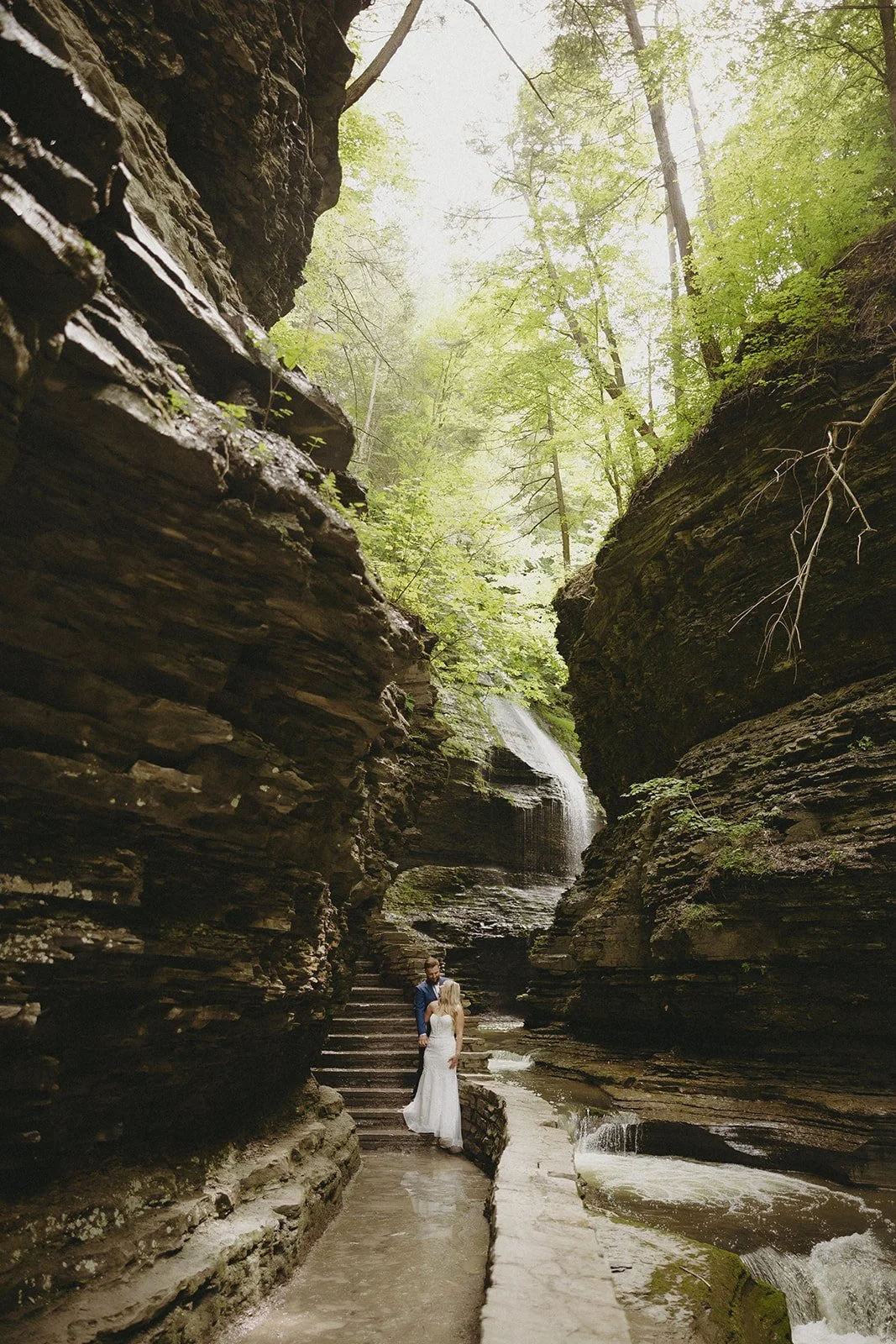 A couple in wedding attire stands by a stone wall in a rocky canyon with a waterfall and lush greenery in the background for Watkins Glen State Park Wedding Photos
