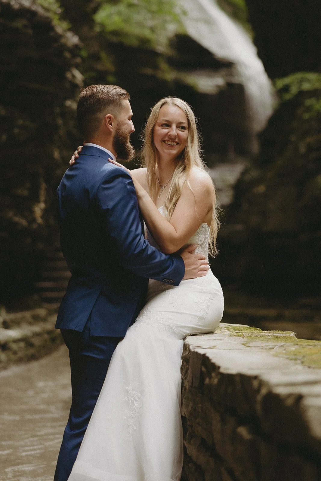 A bride in a white dress sits on a stone ledge while a groom in a blue suit stands in front of her, both smiling outdoors near rocks and a waterfall.
