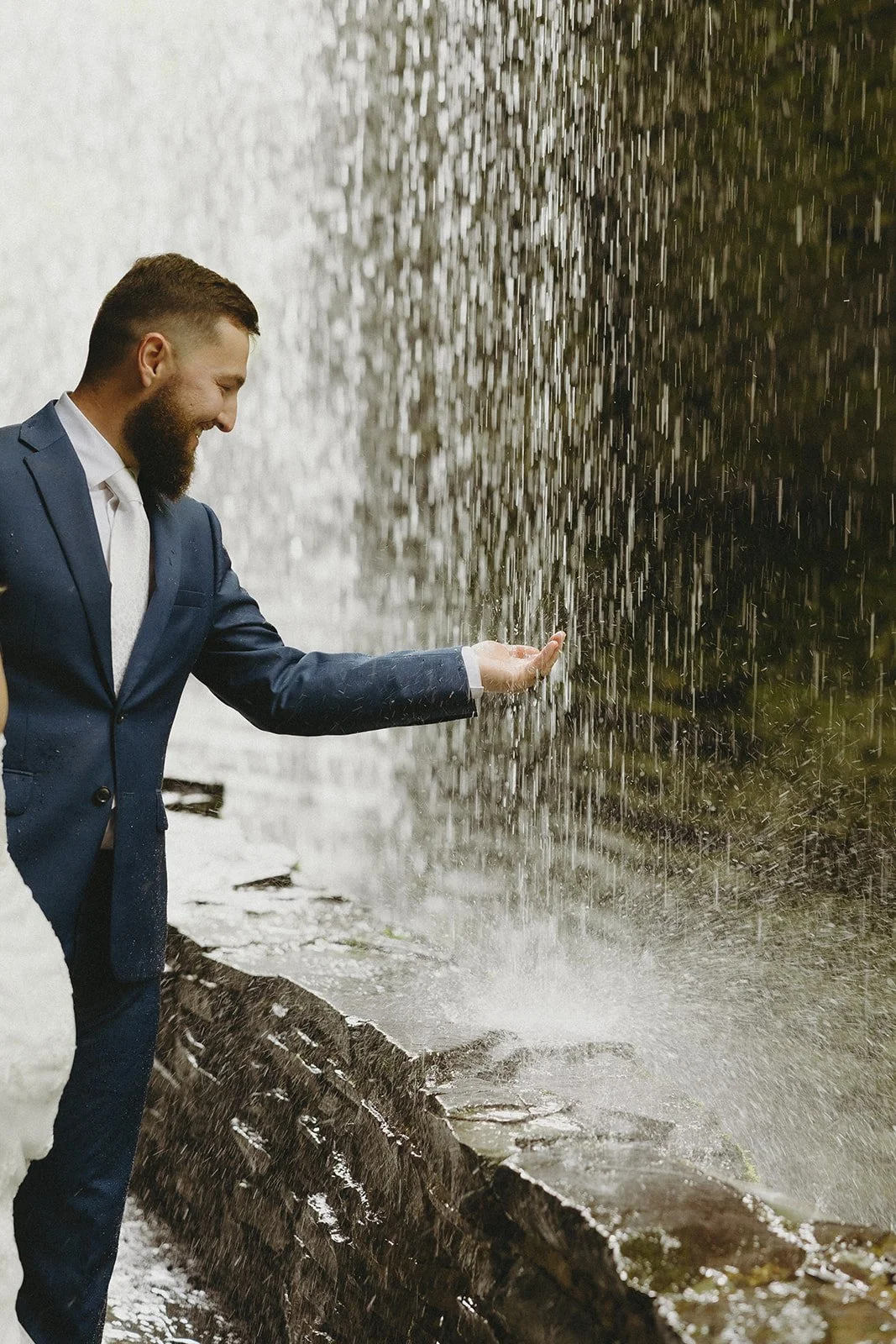 A man in a suit stands by a stone wall, reaching his hand out to touch water falling from a waterfall.