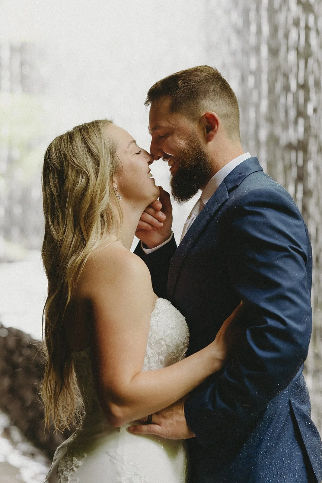 A bride and groom stand close, smiling, and touching noses in front of a waterfall backdrop on their wedding day.