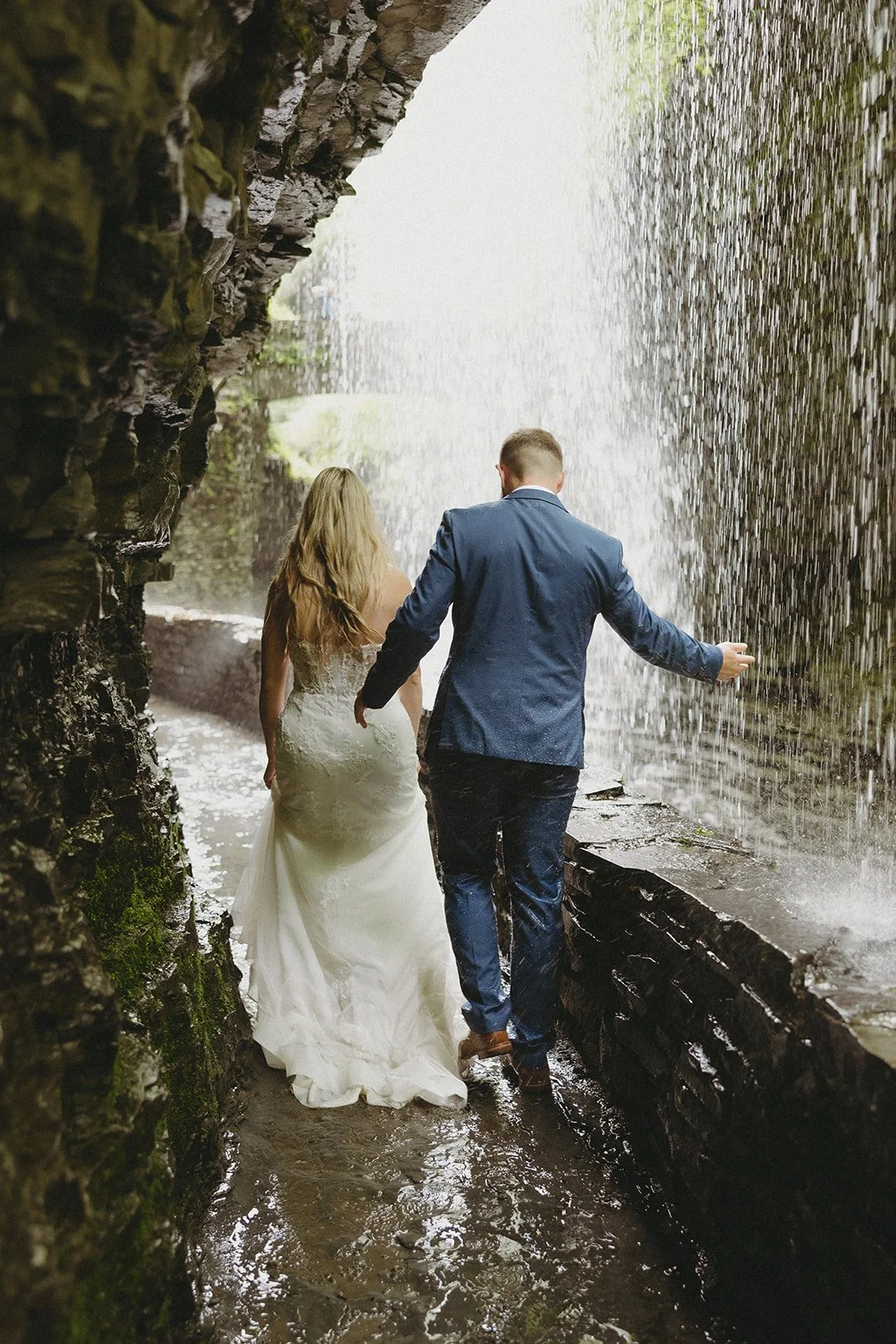 A couple in wedding attire stands by a stone wall in a rocky canyon with a waterfall and lush greenery in the background for Watkins Glen State Park Wedding Photos