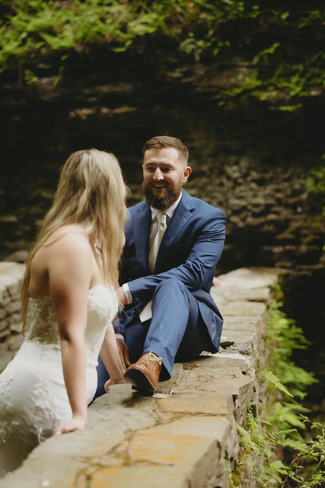 A man in a blue suit and a woman in a white dress sit together on a stone ledge outdoors, surrounded by greenery.