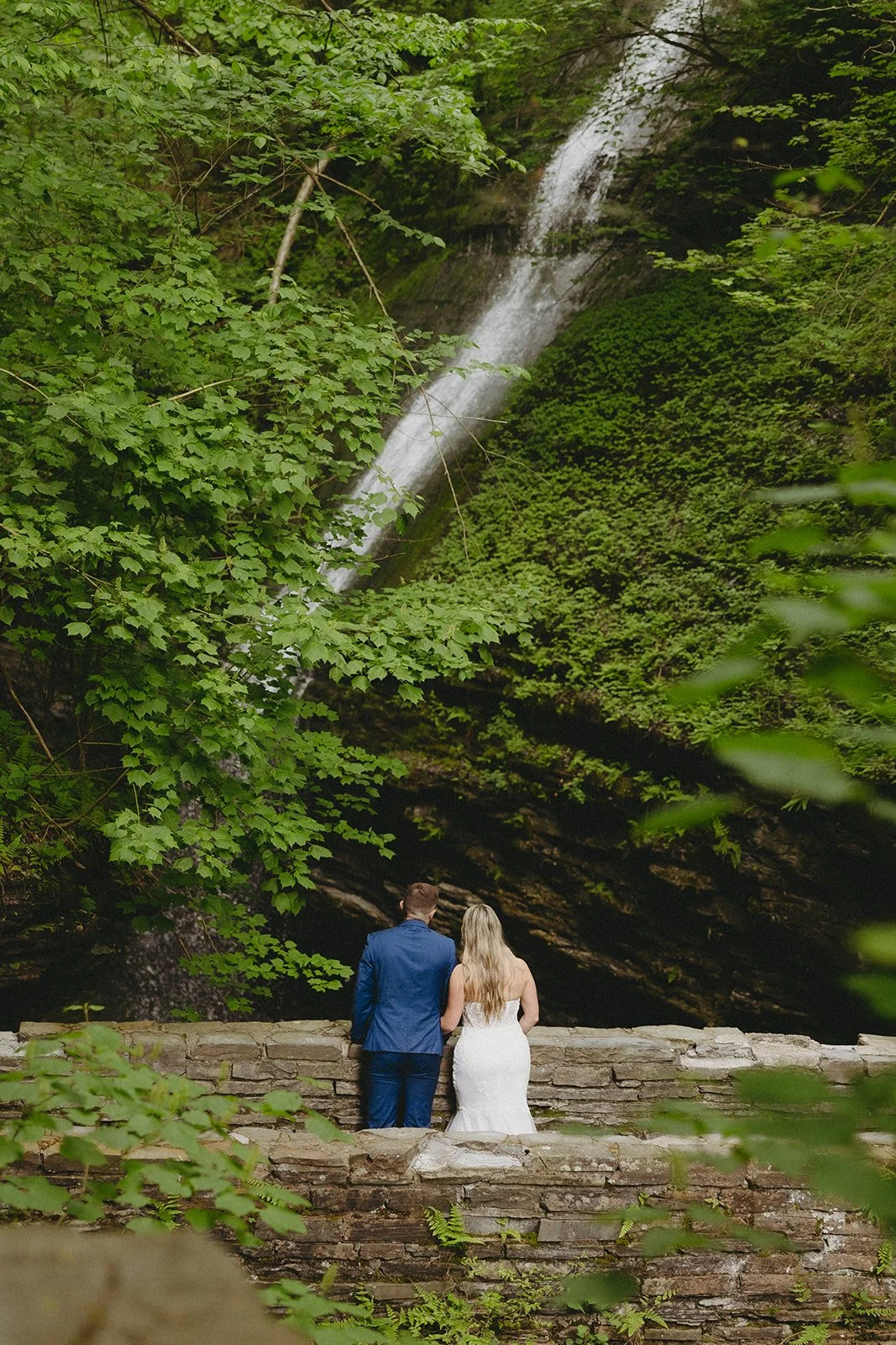 A couple in wedding attire stands by a stone wall in a rocky canyon with a waterfall and lush greenery in the background for Watkins Glen State Park Wedding Photos