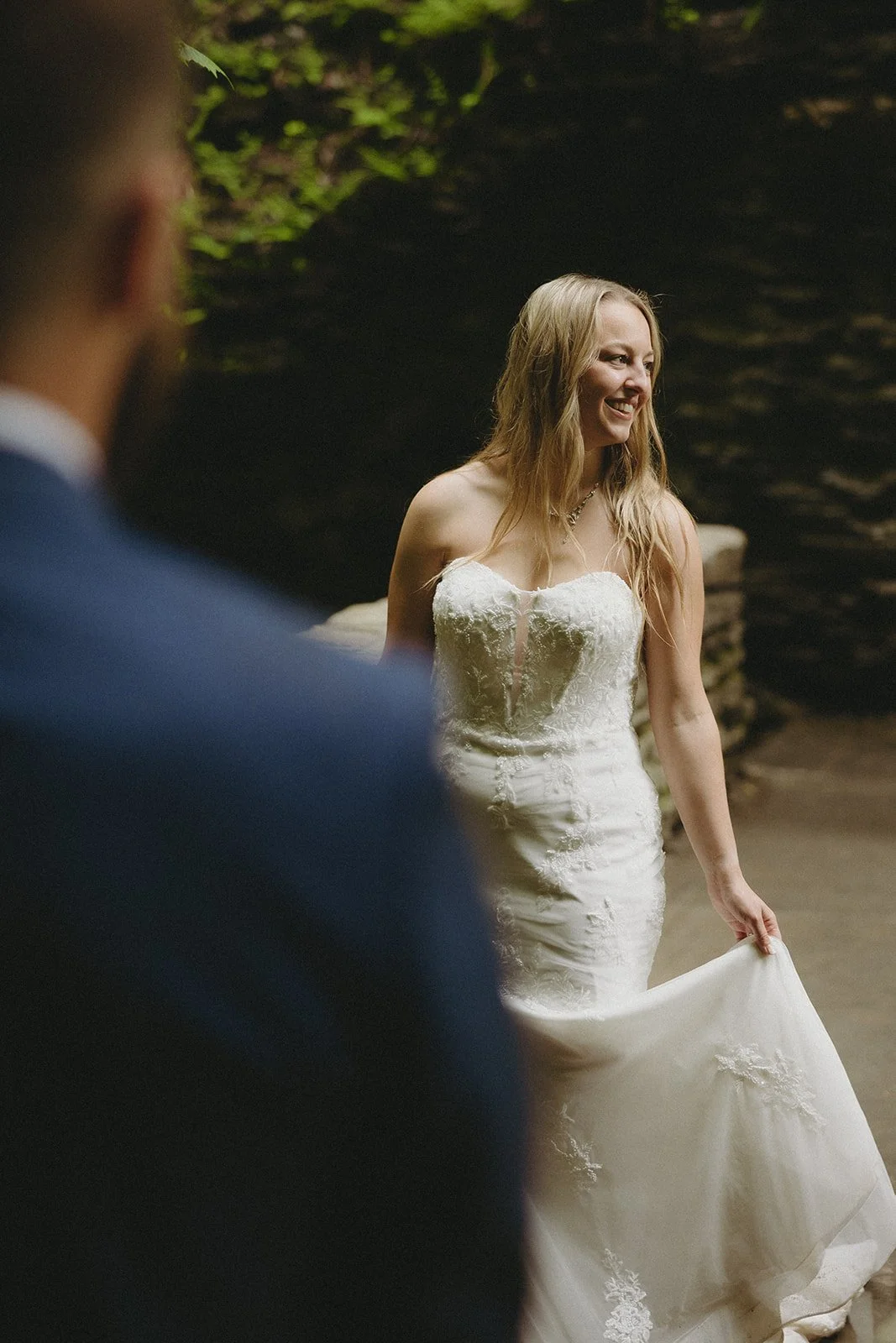 A woman in a strapless white wedding dress smiles and holds her gown while a man in a blue suit stands in the foreground, slightly out of focus.