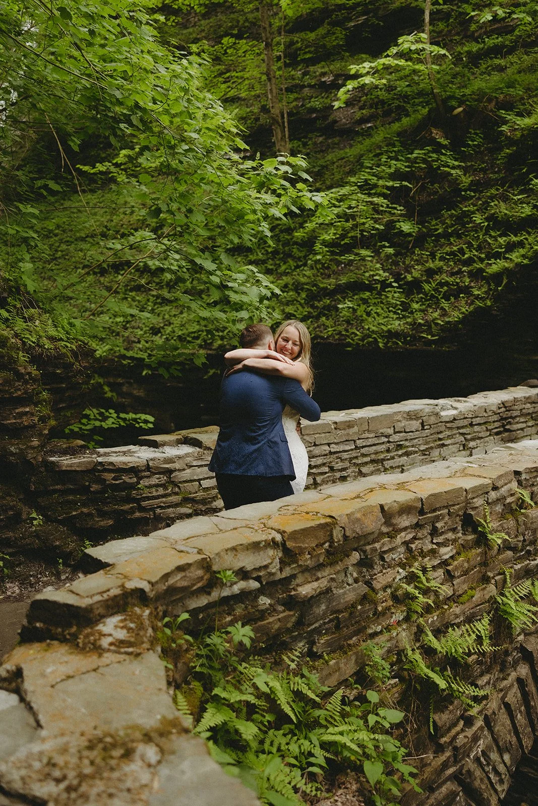 A couple embraces on a stone walkway surrounded by lush greenery and moss-covered walls in an outdoor setting.