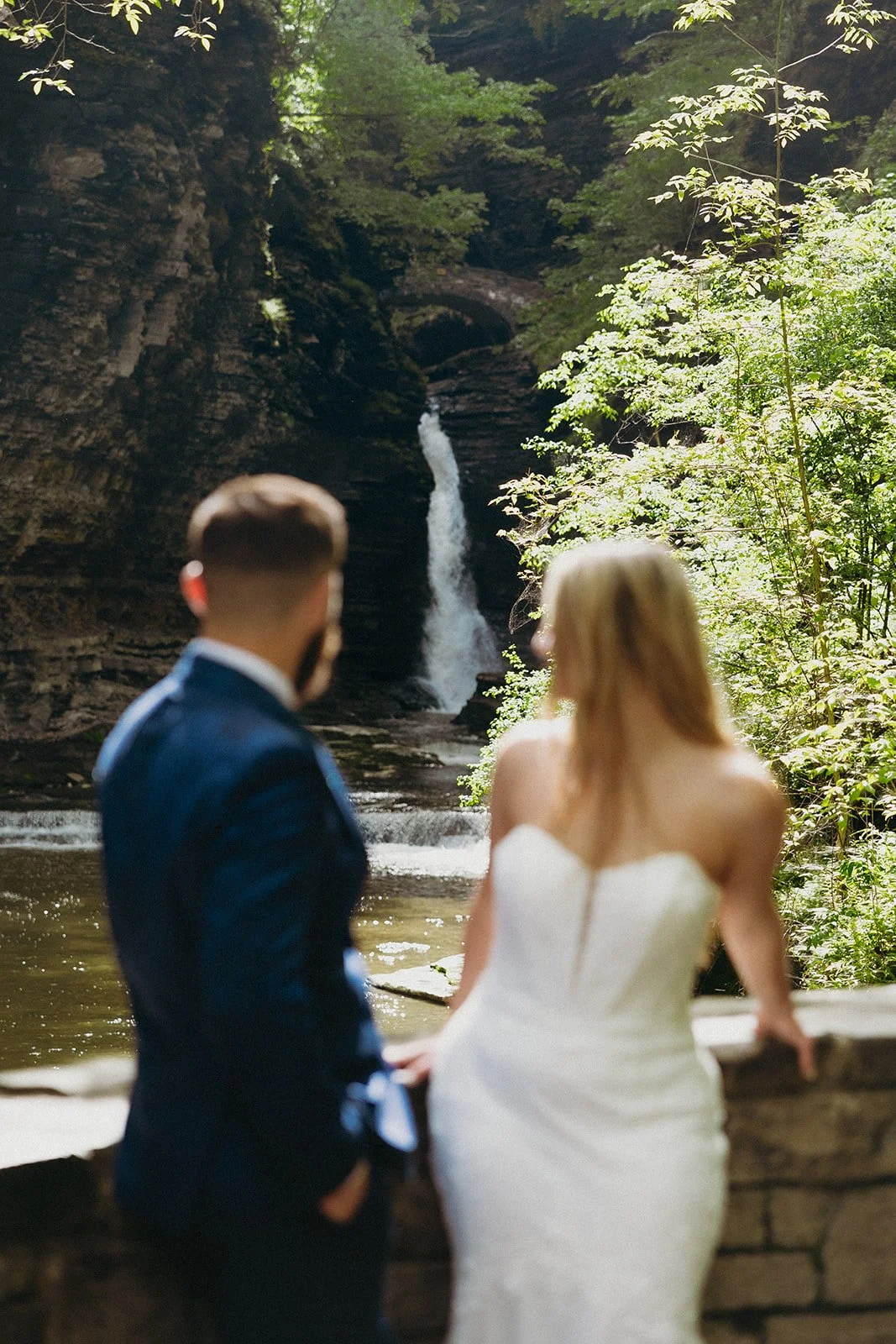 A couple in wedding attire stands by a stone wall in a rocky canyon with a waterfall and lush greenery in the background for Watkins Glen State Park Wedding Photos