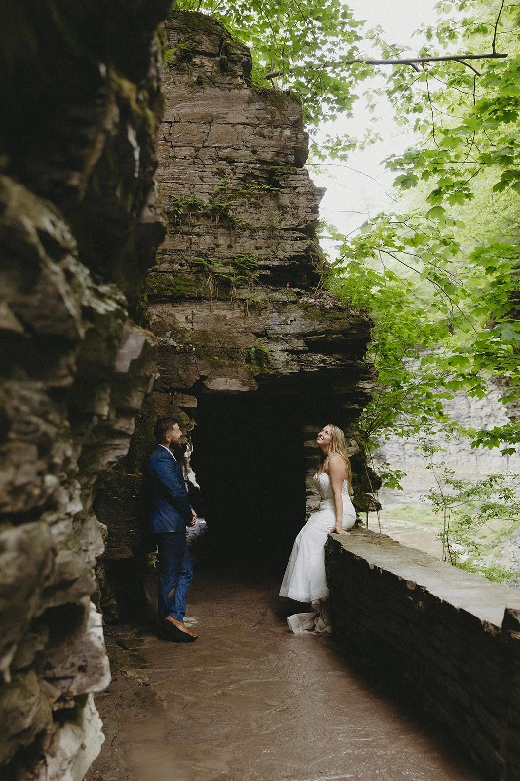 A bride and groom stand facing each other near a rocky tunnel surrounded by greenery.