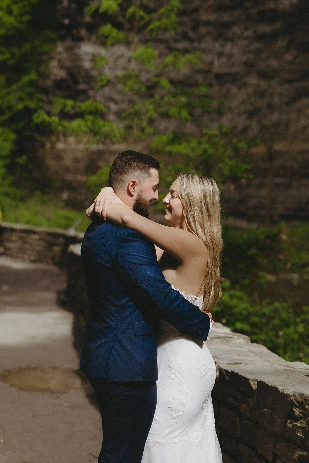 A couple in formal attire embraces outdoors by a stone wall, with greenery and a rocky background visible.