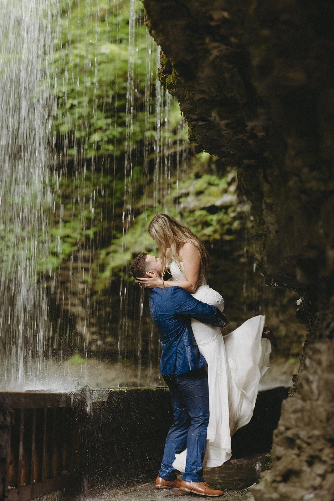 A couple in wedding attire stands by a stone wall in a rocky canyon with a waterfall and lush greenery in the background for Watkins Glen State Park Wedding Photos