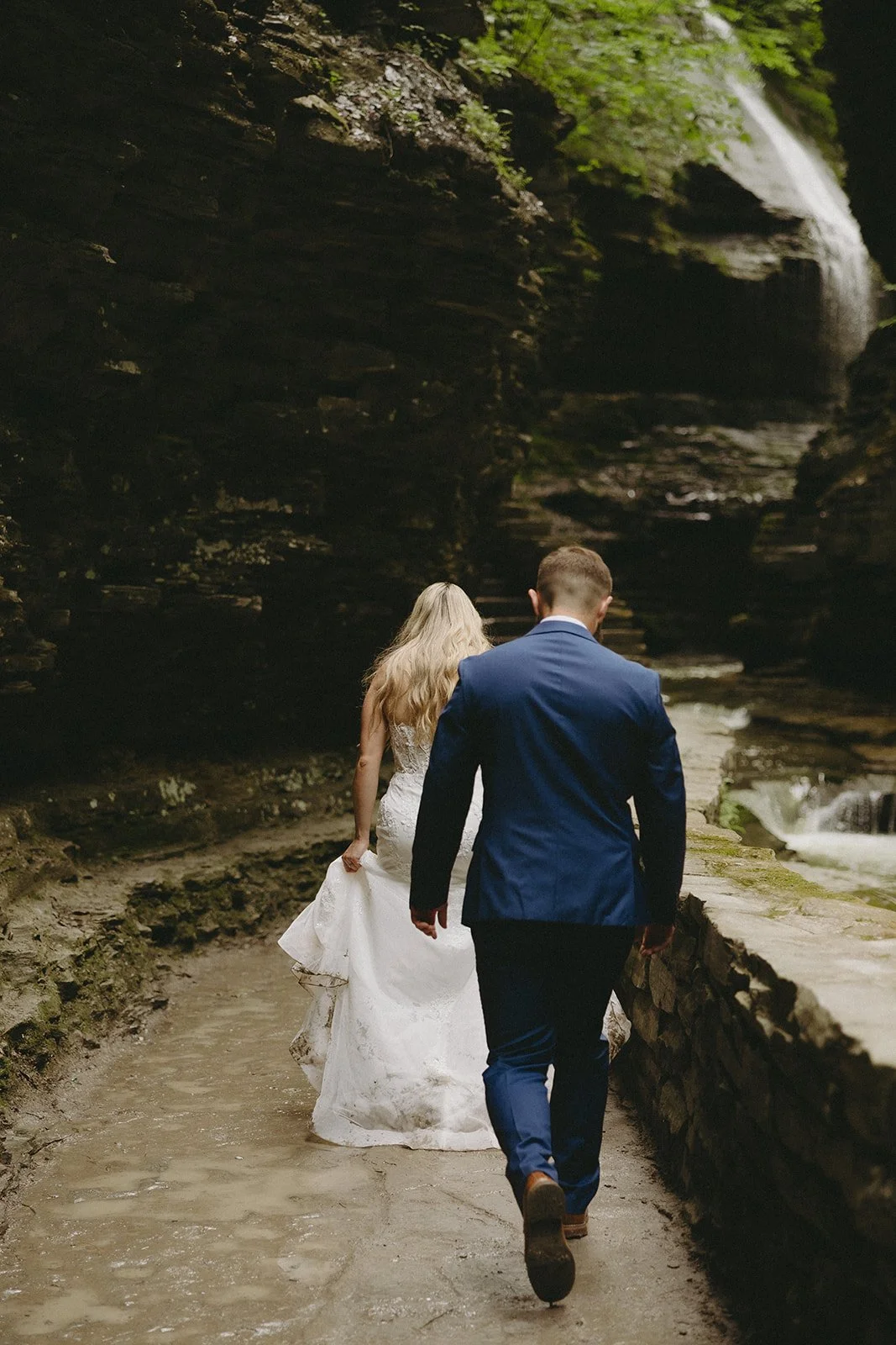 A couple dressed in wedding attire walks along a rocky path beside a stream in a wooded area, with a waterfall visible in the background.