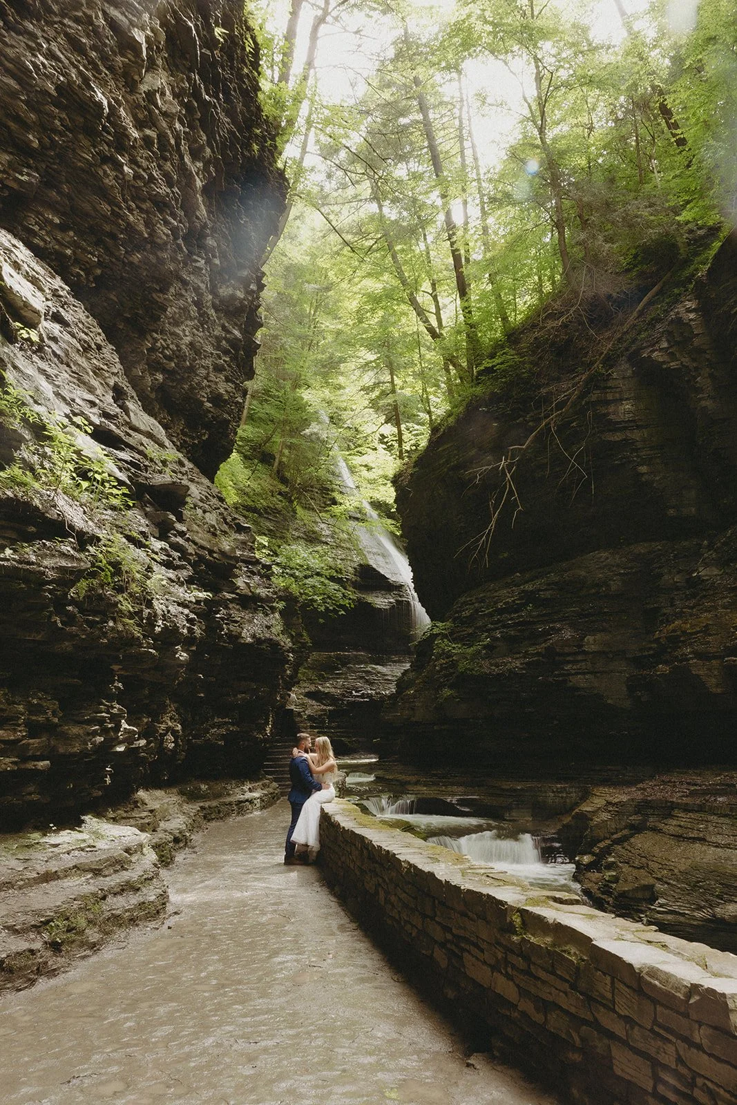 A couple in wedding attire stands by a stone wall in a rocky canyon with a waterfall and lush greenery in the background for Watkins Glen State Park Wedding Photos