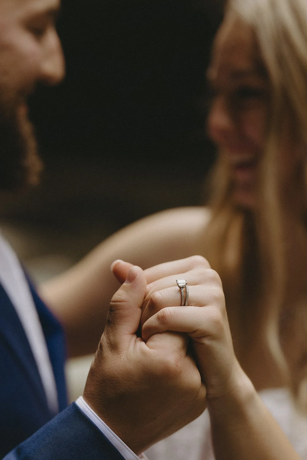 Close-up of a couple holding hands, with a focus on the woman’s engagement ring; both are smiling and dressed formally.