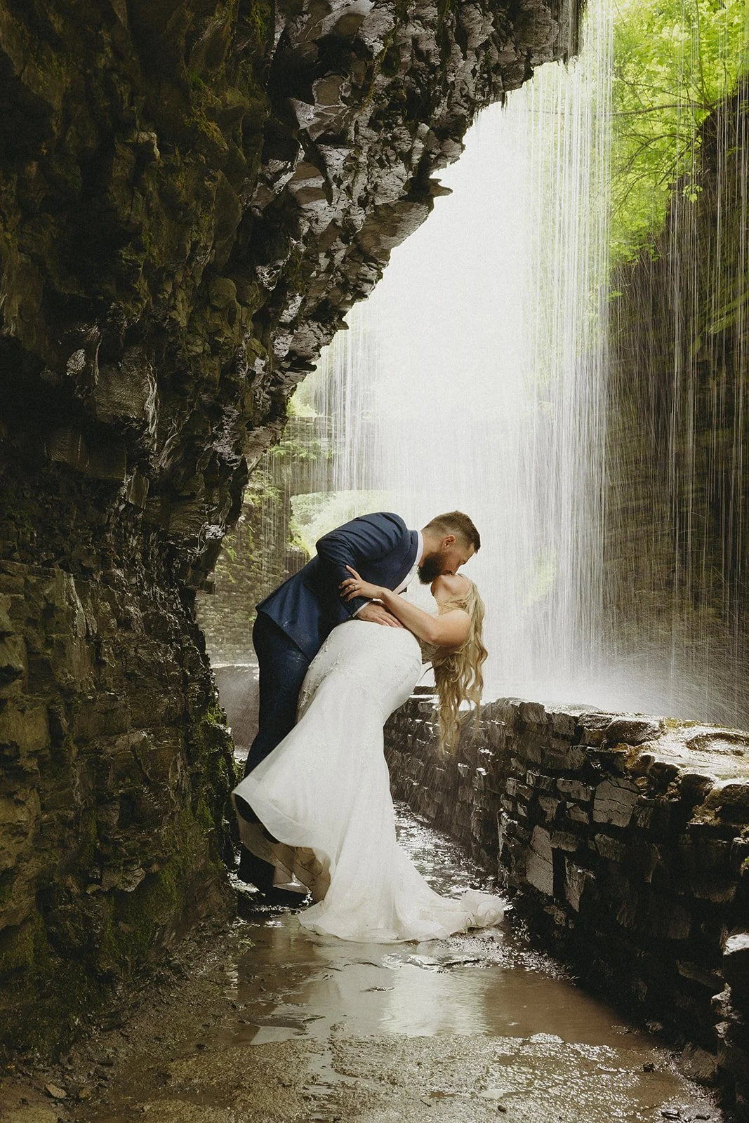 A couple in wedding attire stands by a stone wall in a rocky canyon with a waterfall and lush greenery in the background for Watkins Glen State Park Wedding Photos