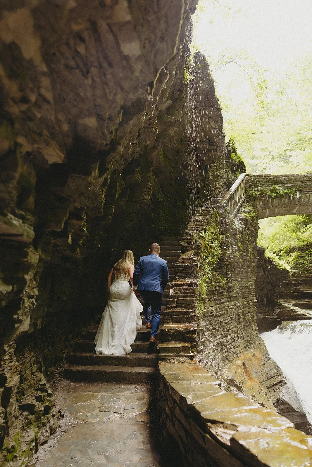 A bride and groom walk up stone steps along a rocky, moss-covered path beside a waterfall in a natural outdoor setting.