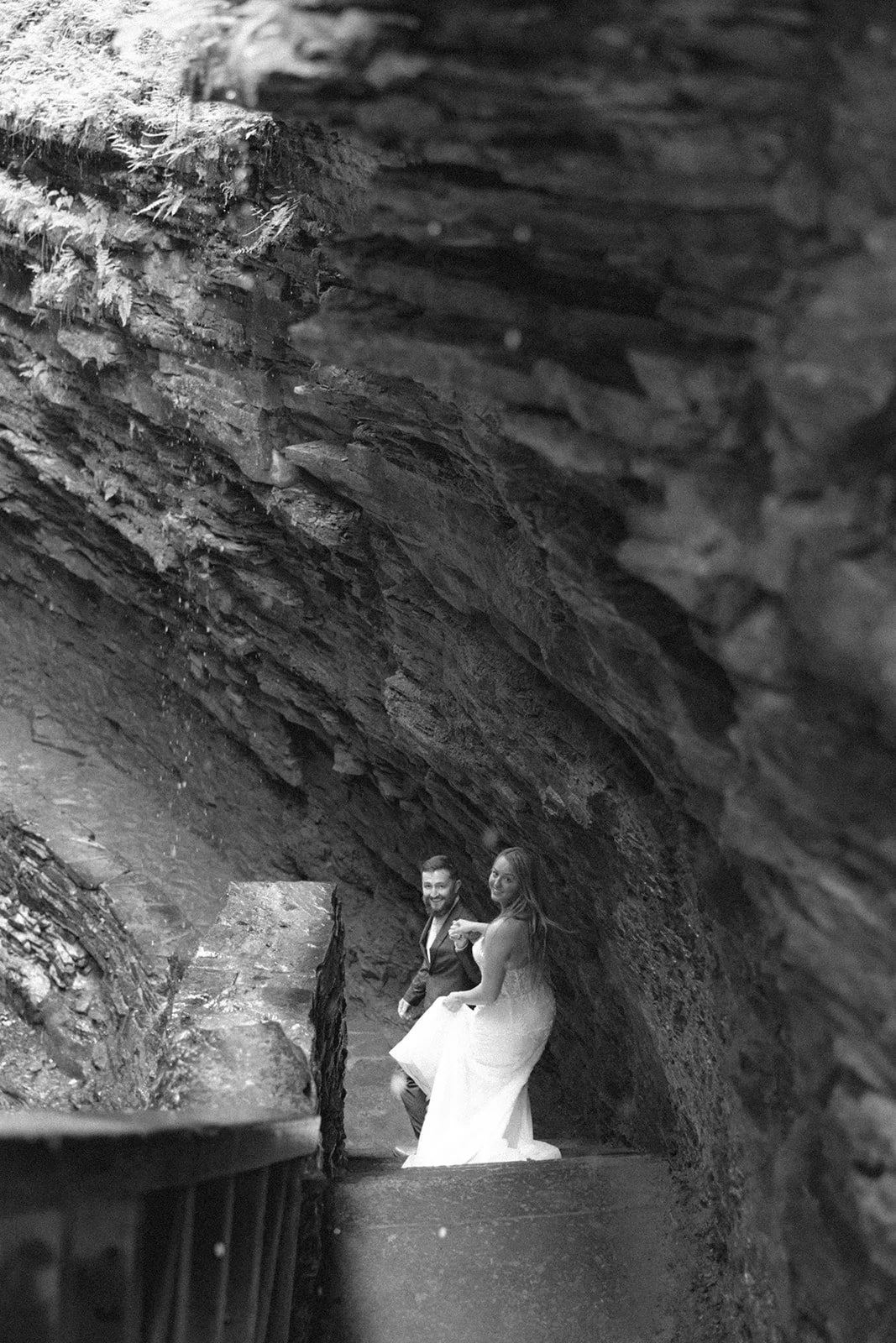 A couple in wedding attire stands by a stone wall in a rocky canyon with a waterfall and lush greenery in the background for Watkins Glen State Park Wedding Photos