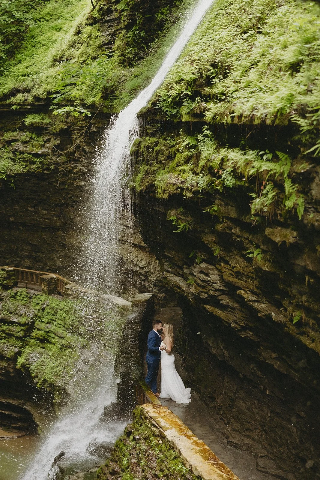A bride and groom stand together beside a tall waterfall surrounded by moss-covered rock walls and greenery.