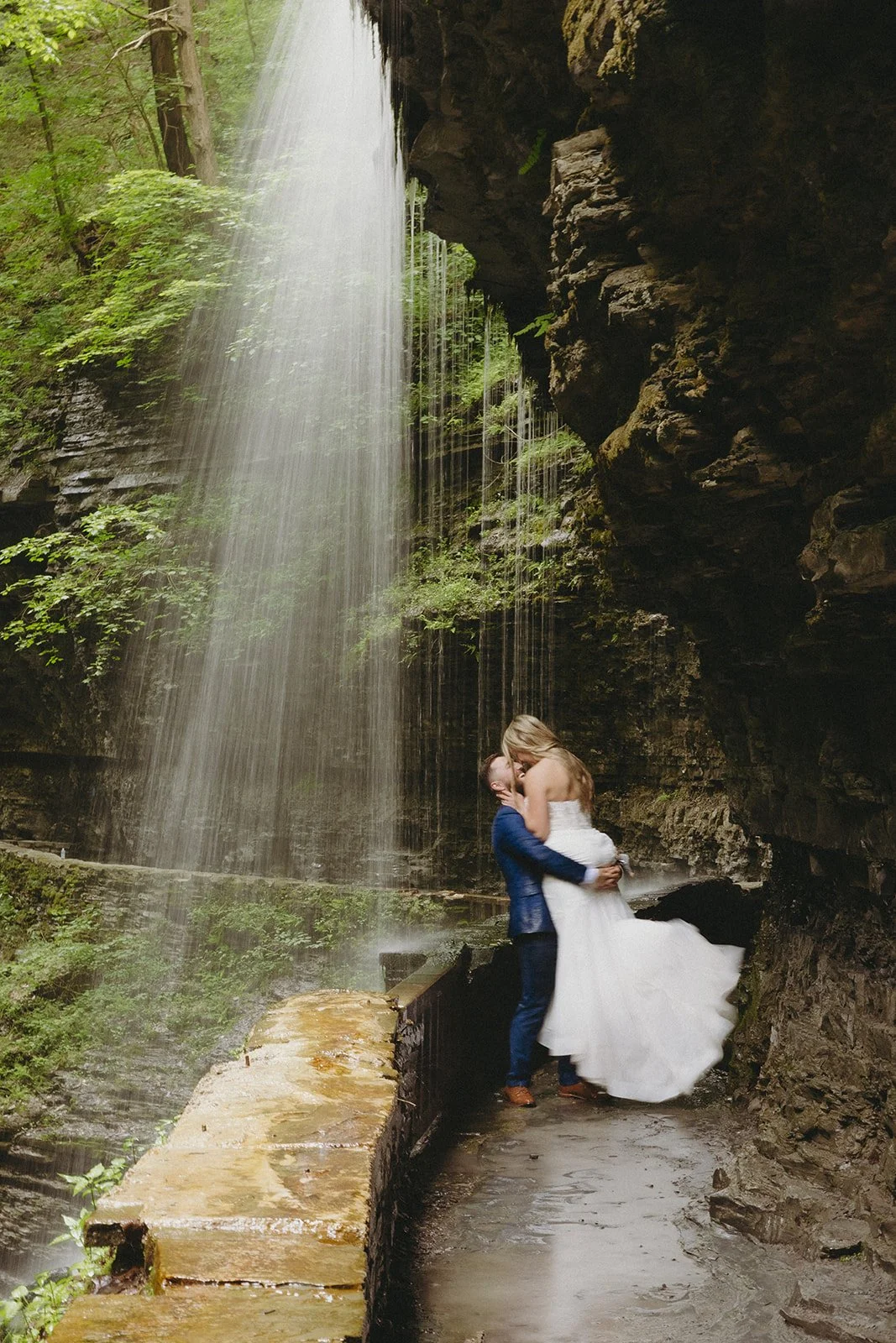 A bride and groom stand together beside a tall waterfall surrounded by moss-covered rock walls and greenery.