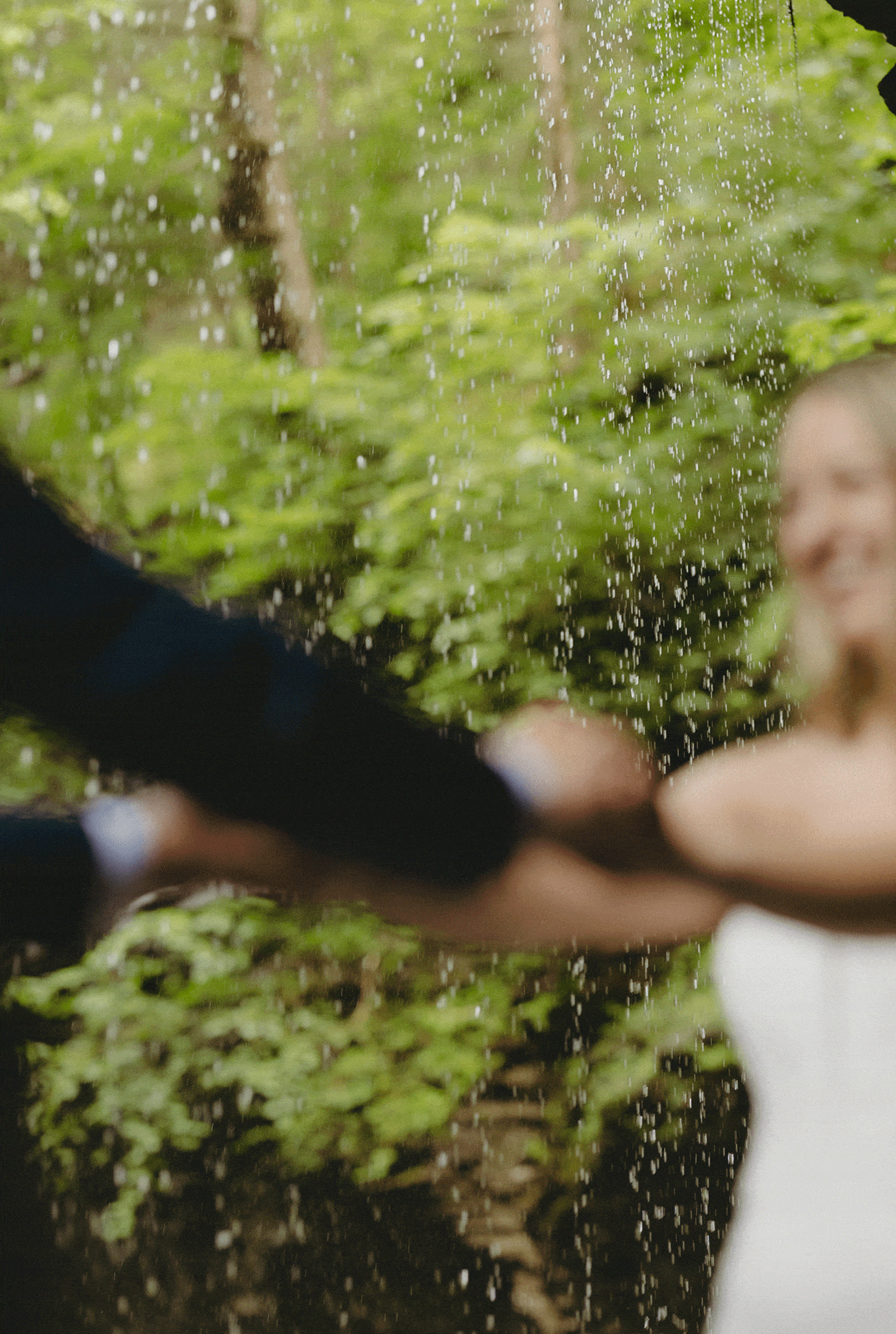 A bride and groom hold hands and spin around in front of a curtain of falling water, with a blurred forest background.